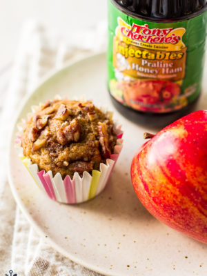 An apple praline muffin on a plate with an apple and a bottle of syrup