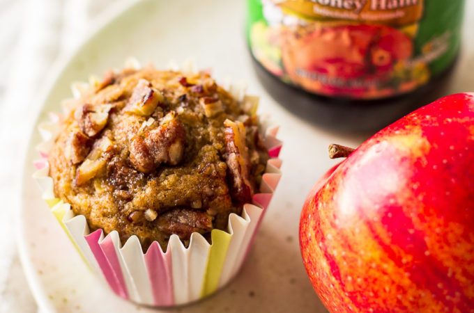 An apple praline muffin on a plate with an apple and a bottle of syrup