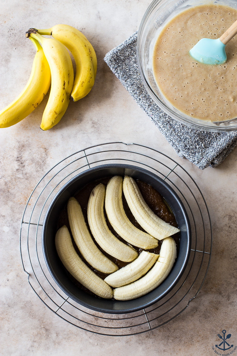 Overhead photo of a prebaked banana upside down cake with three bananas and a bowl of batter