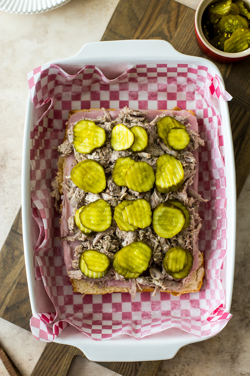 Overhead photo of pickle topped sliders in a baking dish with red and white checked parchment