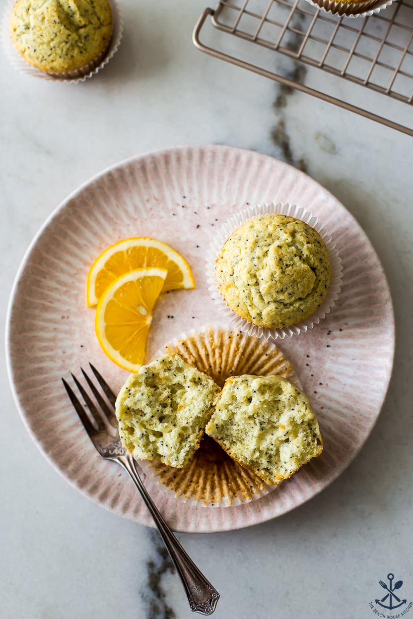 Overhead photo of a split open lemon poppy seed muffin on a pink plate with another muffins to two slices of lemon