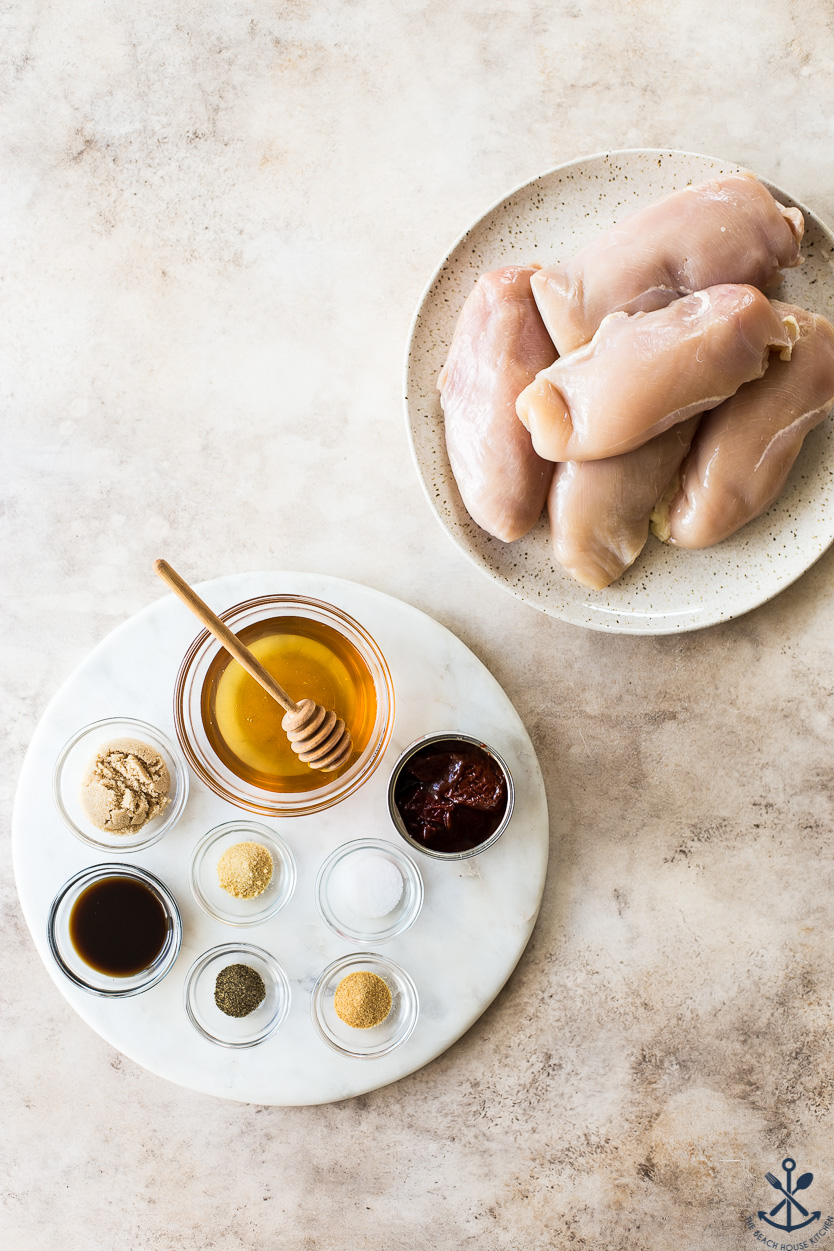 Overhead photo of a plate of uncooked chicken breads and ingredients for a honey sauce on a round marble board