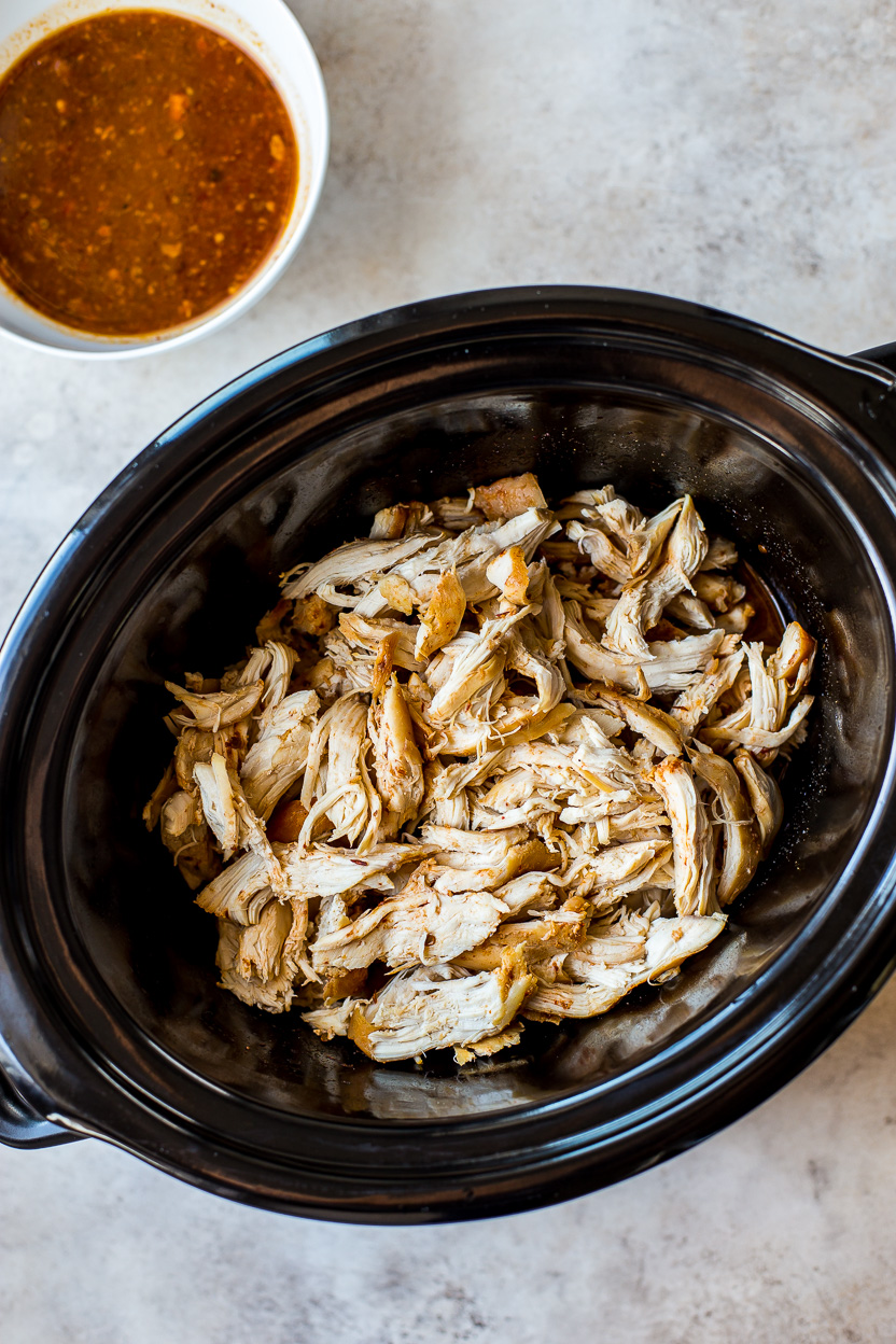 Overhead photo of shredded chicken in a slow cooker with a bowl of sauce off to the side