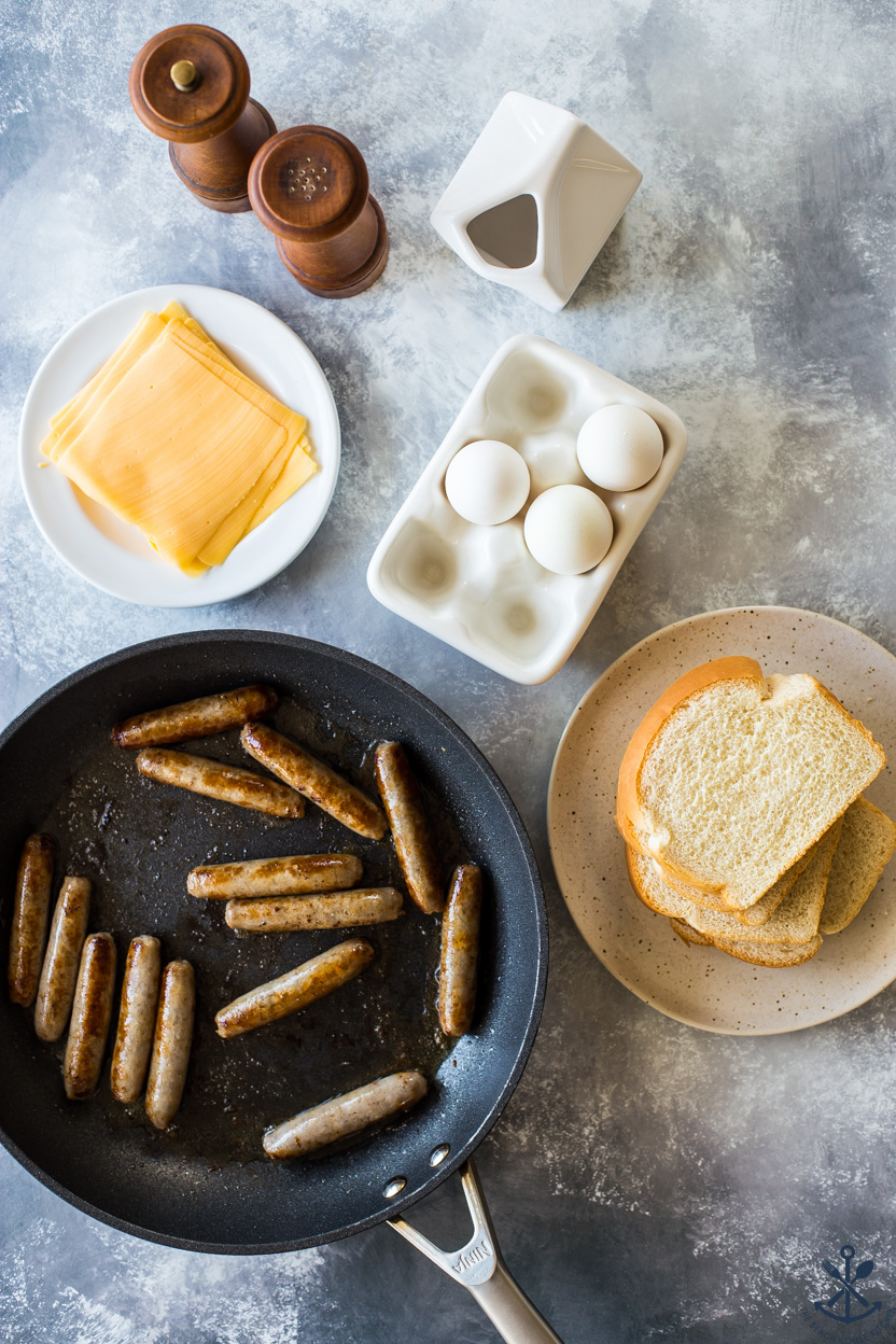 Overhead photo of a skillet of sausage links, a plate of cheese slices, eggs, bread, milk and salt and pepper