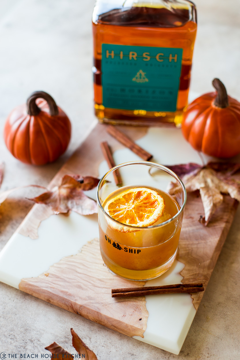 A pumpkin spice old fashioned cocktail on a board with a bottle of bourbon, pumpkins, leaves and cinnamon sticks