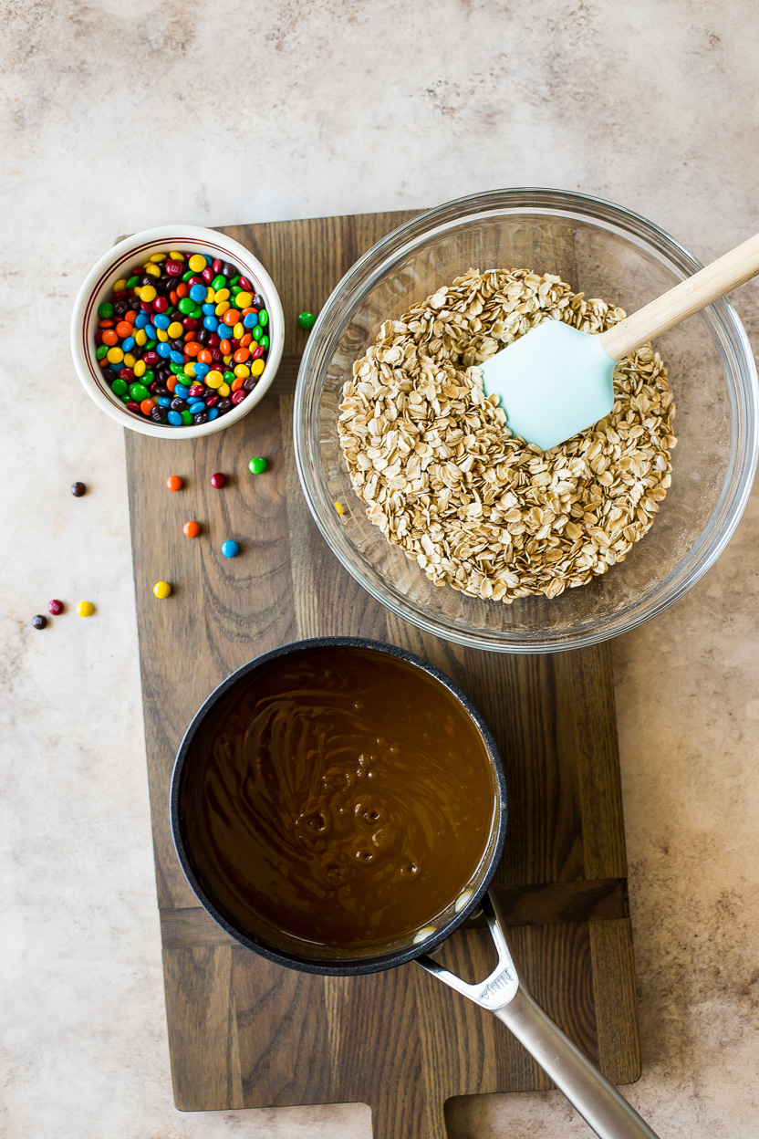Overhead photo of a bowl of oats, a bowl of M&M's and a sauce pan with a brown mixture