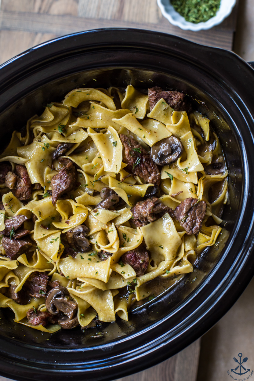 Up close overhead photo of a black slow cooker of noodles with beef and mushrooms
