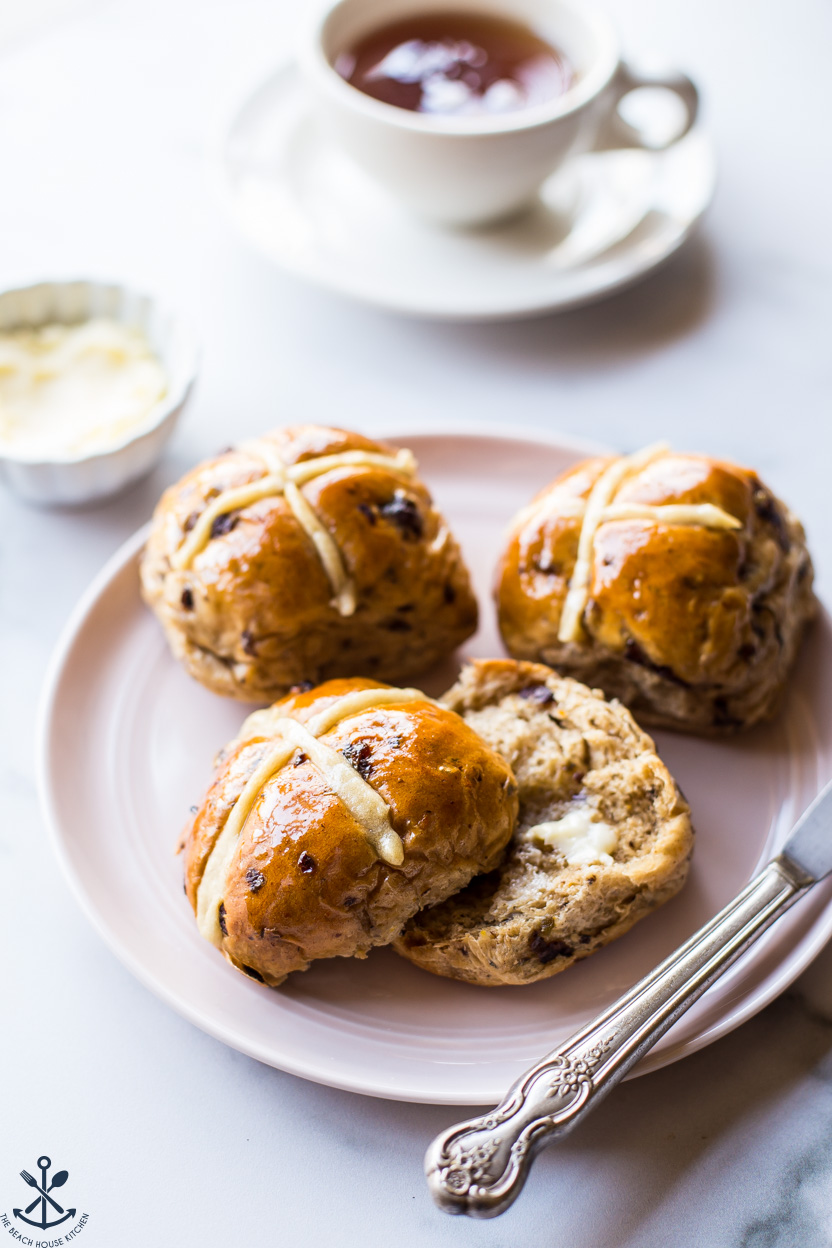 Up close photo of a plate of hot cross buns with a cup of tea in the background