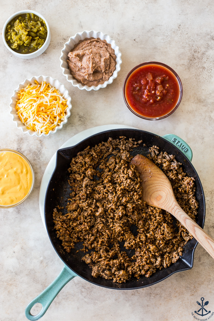 Overhead photo of a skillet of cooked taco meat surrounded by some bowls of cheese, salsa, refried beans and green chiles