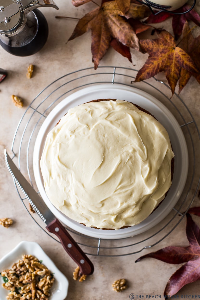 Overhead photo of a cream cheese frosted maple walnut cake on a plate on a round wire rack surrounded by fall leaves