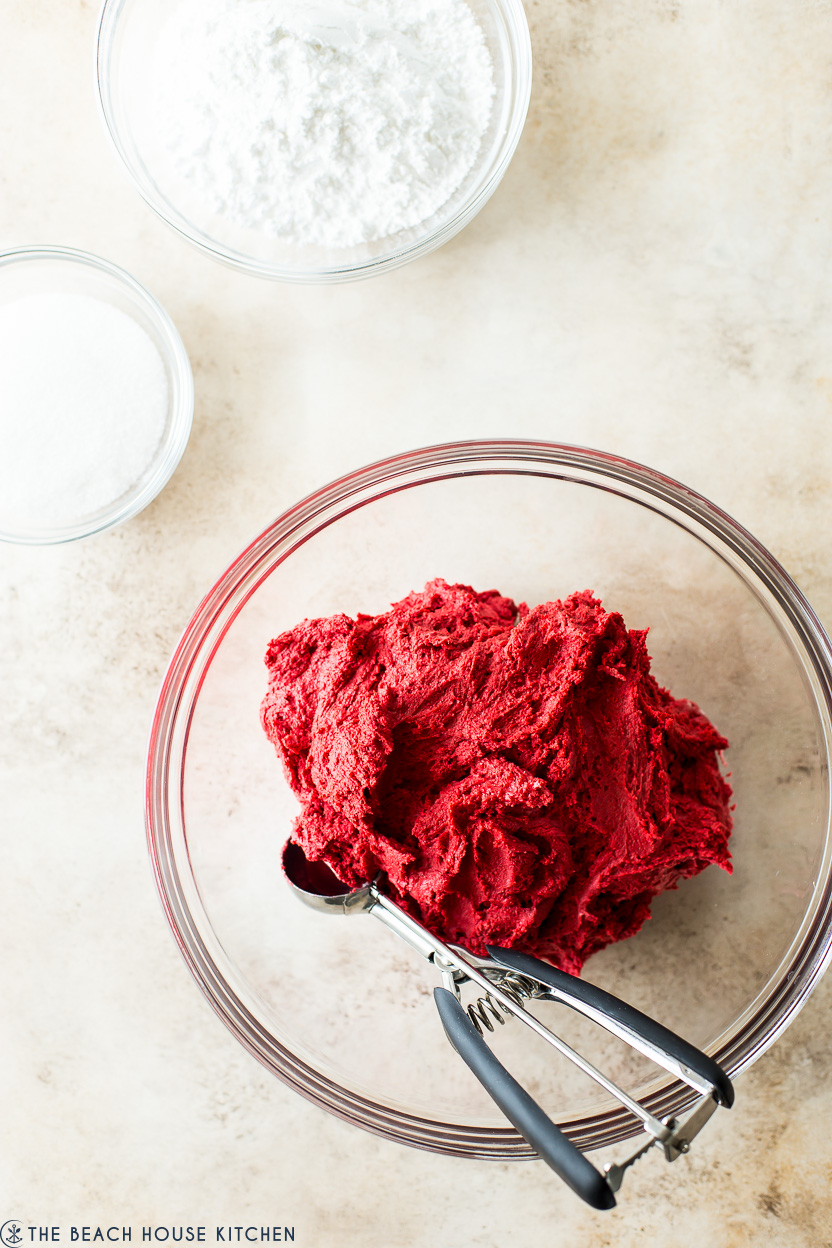 Overhead photo of a bowl of red velvet cookie dough, a bowl of granulated sugar and a bowl of confectioners' sugar