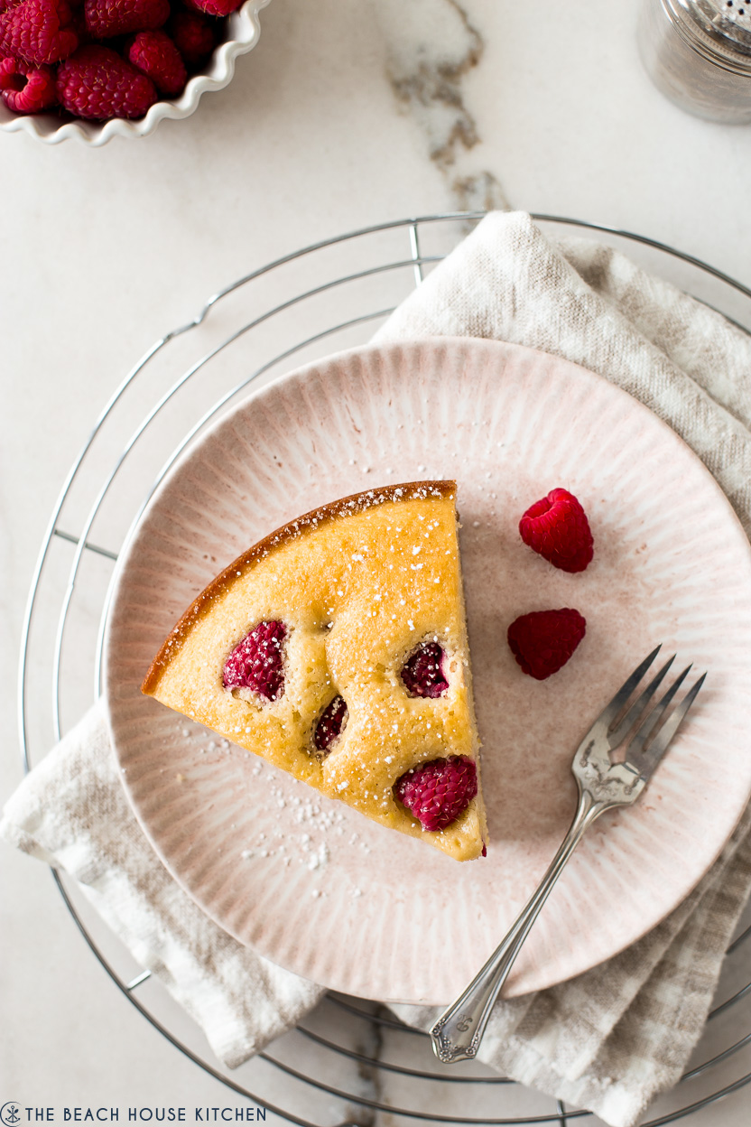 Overhead photo of a slice of cake topped with fresh raspberries on a pink plate with a fork and a few raspberries