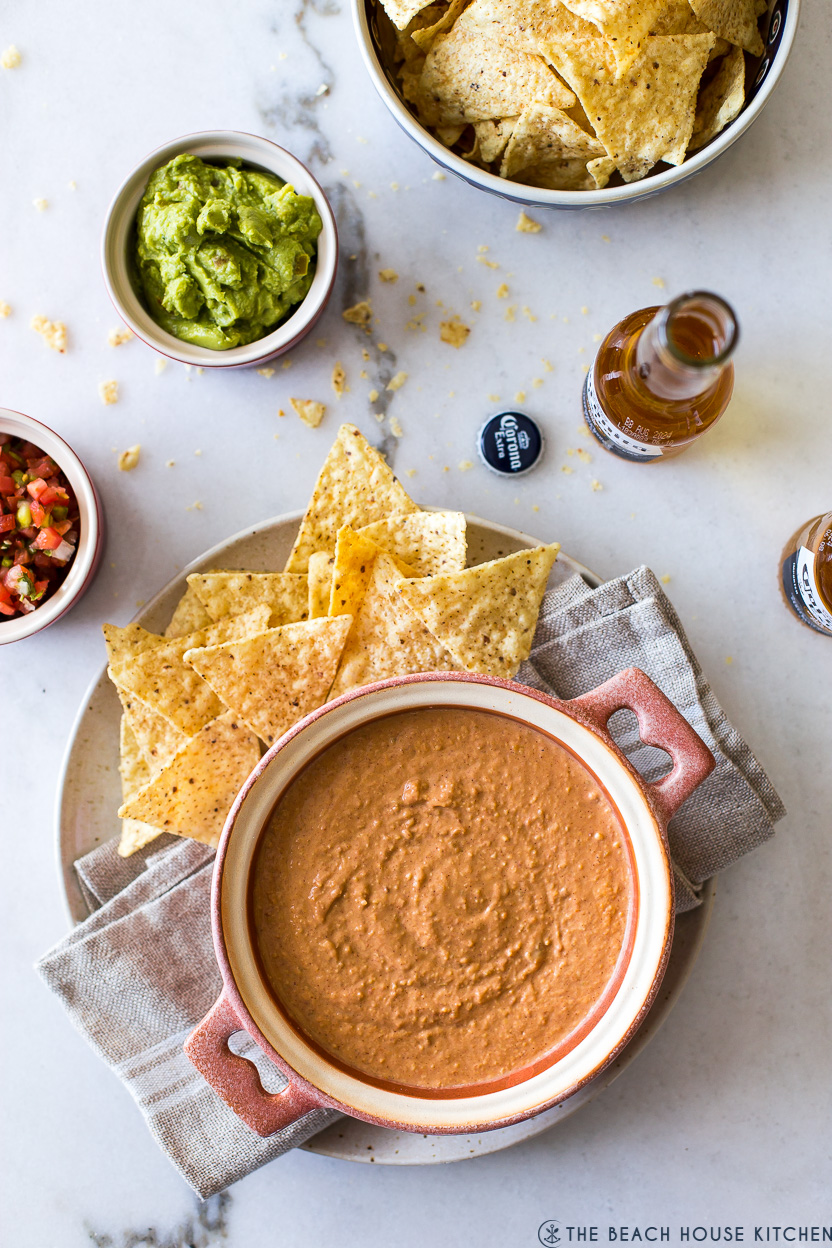 Overhead photo of a bean dip in a baking dish with tortillas and a bowl of guacamole, beers and a bowl of tortilla chips