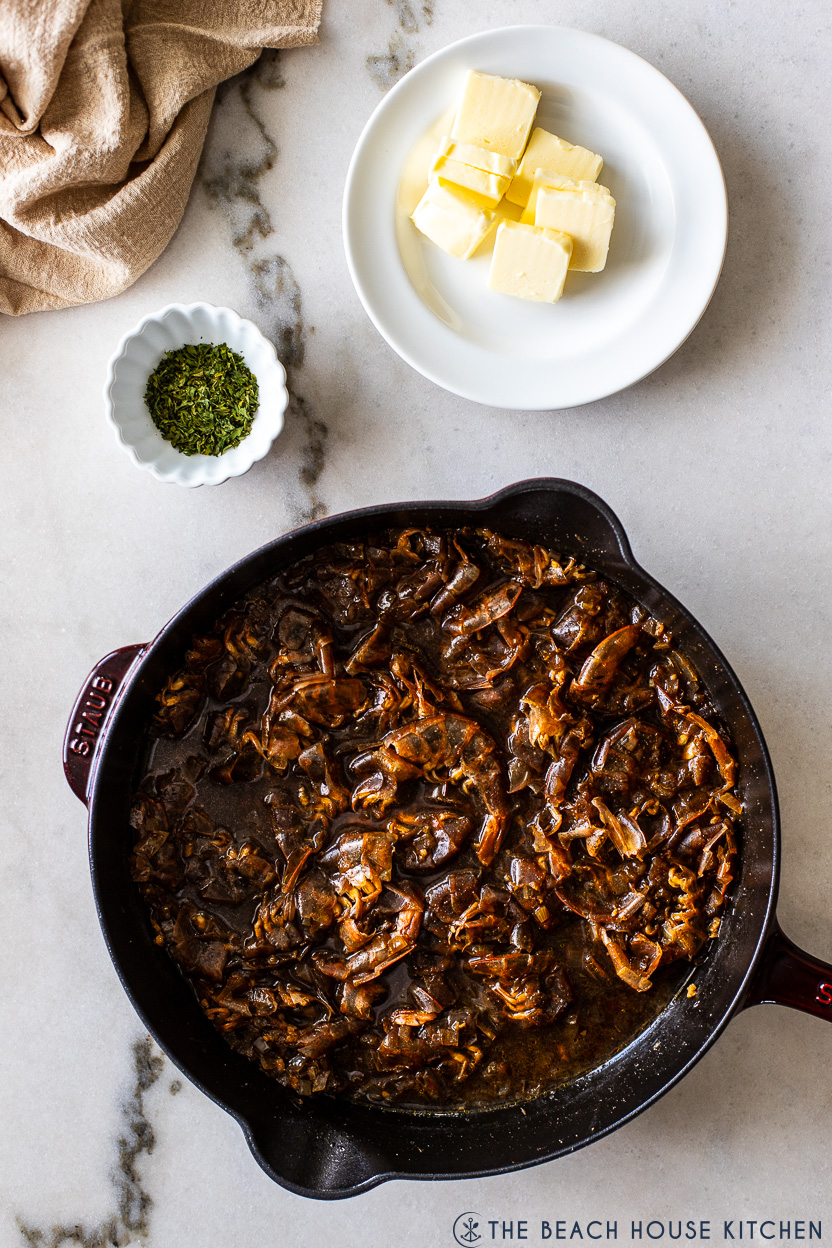 Overhead photo of a skillet filled with shrimp shells and a bbq base with a plate of butter patties and a small bowl of chopped parsley