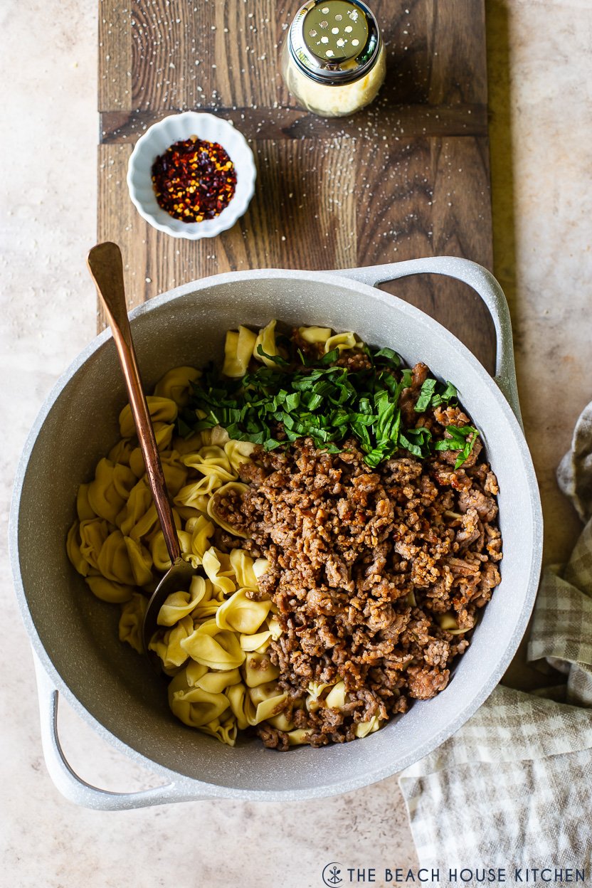 Overhead photo of a large pot filled with cooked Italian sausage, tortellini and basil