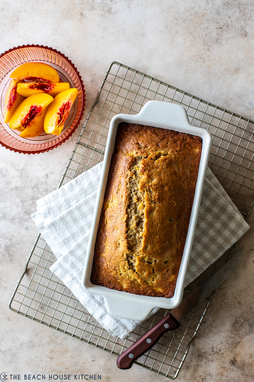 Overhead photo of a loaf of baked peach bread in a loaf pan on a wire rack with a small bowl of peaches