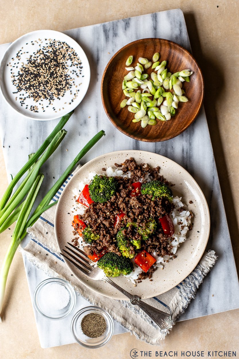 Overhead photo of a plate of Korean ground beef with broccoli and peppers on a marble board with a plate of sliced green onions and a plate of sesame seeds