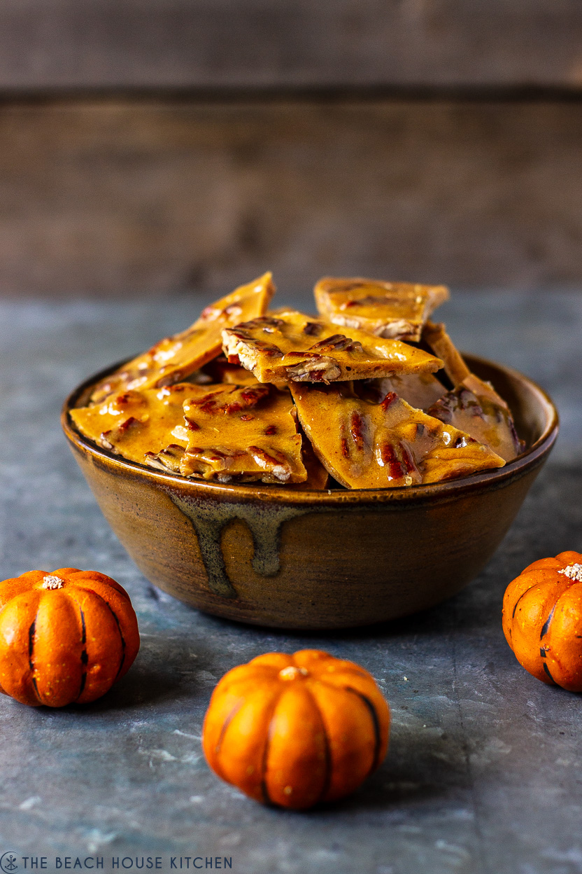 A bowl of pumpkin spice pecan brittle with a few little pumpkins surrounding the bowl
