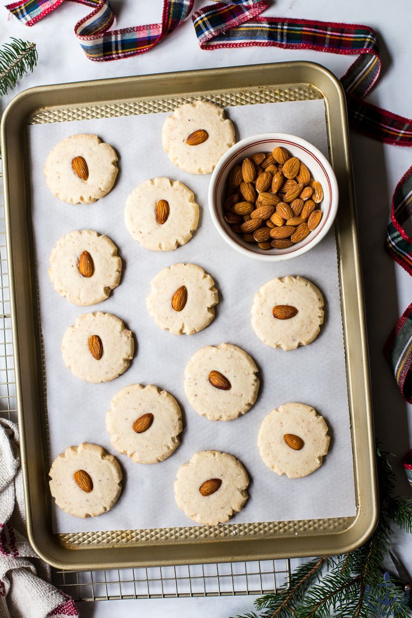 Overhead photo of a baking sheet of almond cookies with a whole almond in the middle of each cookies and a small bowl of whole almonds