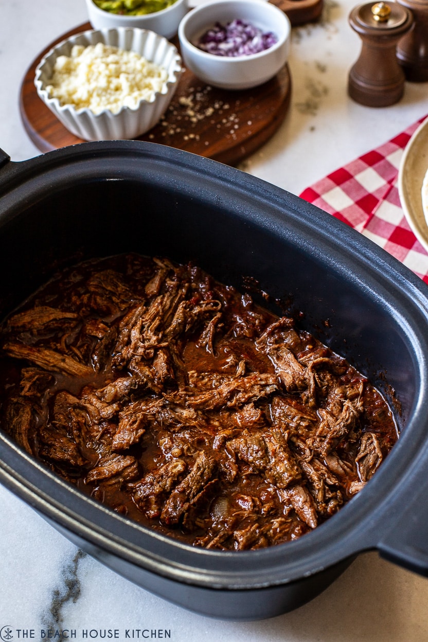 A slow cooker of beef tonga with a few bowls of food in the background