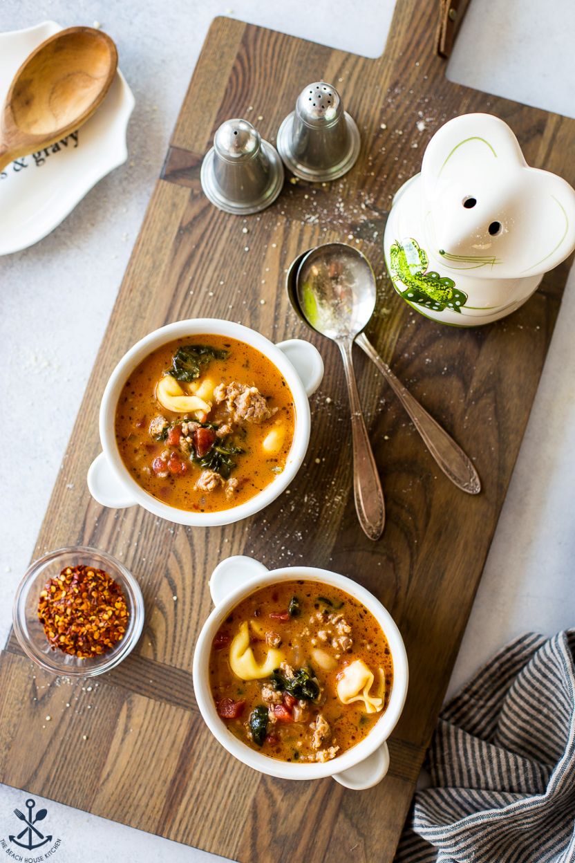 Overhead photo of two bowls of tomato tortellini stew on a wooden board with two spoons, a mouse ceramic cheese conatiner and a silver salt and pepper shaker.