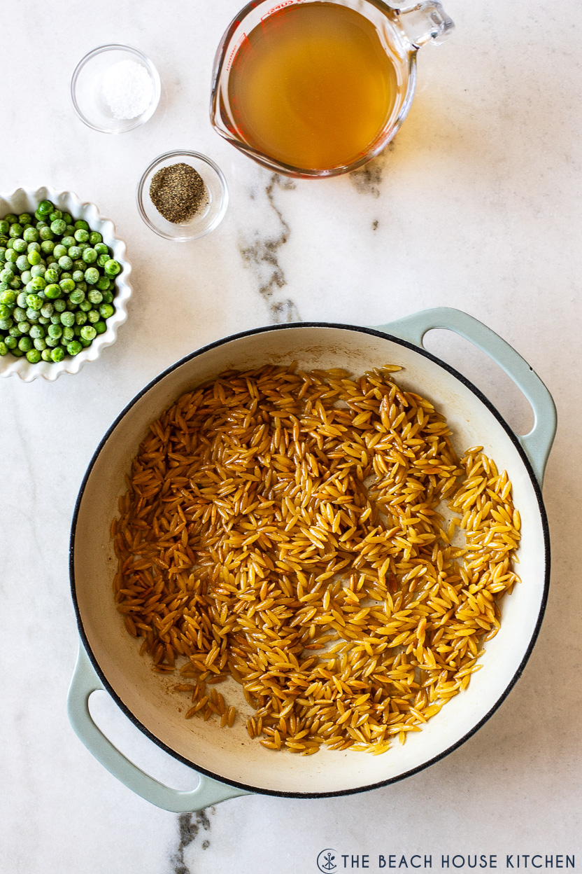 Overhead photo of a skillet of cooked orzo with a measuring cup of broth, a small bowl of pas and salt and pepper off to the top left