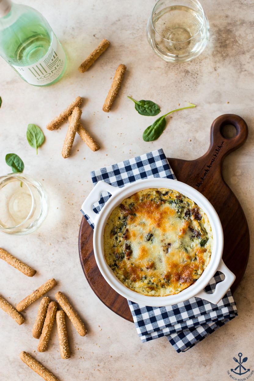 Overhead photo of a small crock of spinach artichoke dip with bacon surrounded by breadsticks and a bottle and glass of wine