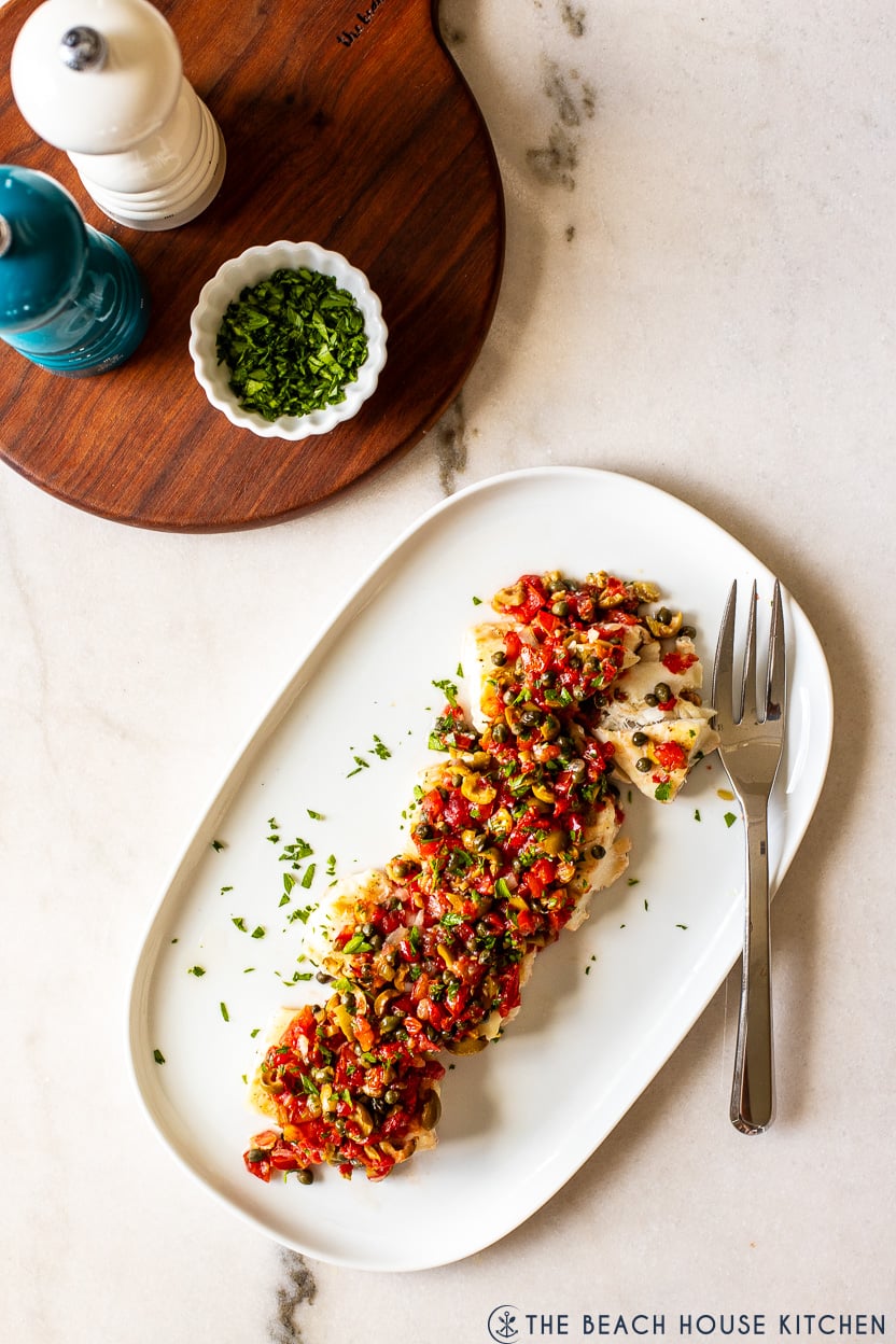 Overhead photo of a white oval platter of baked cod topped with pico de gallo and olives