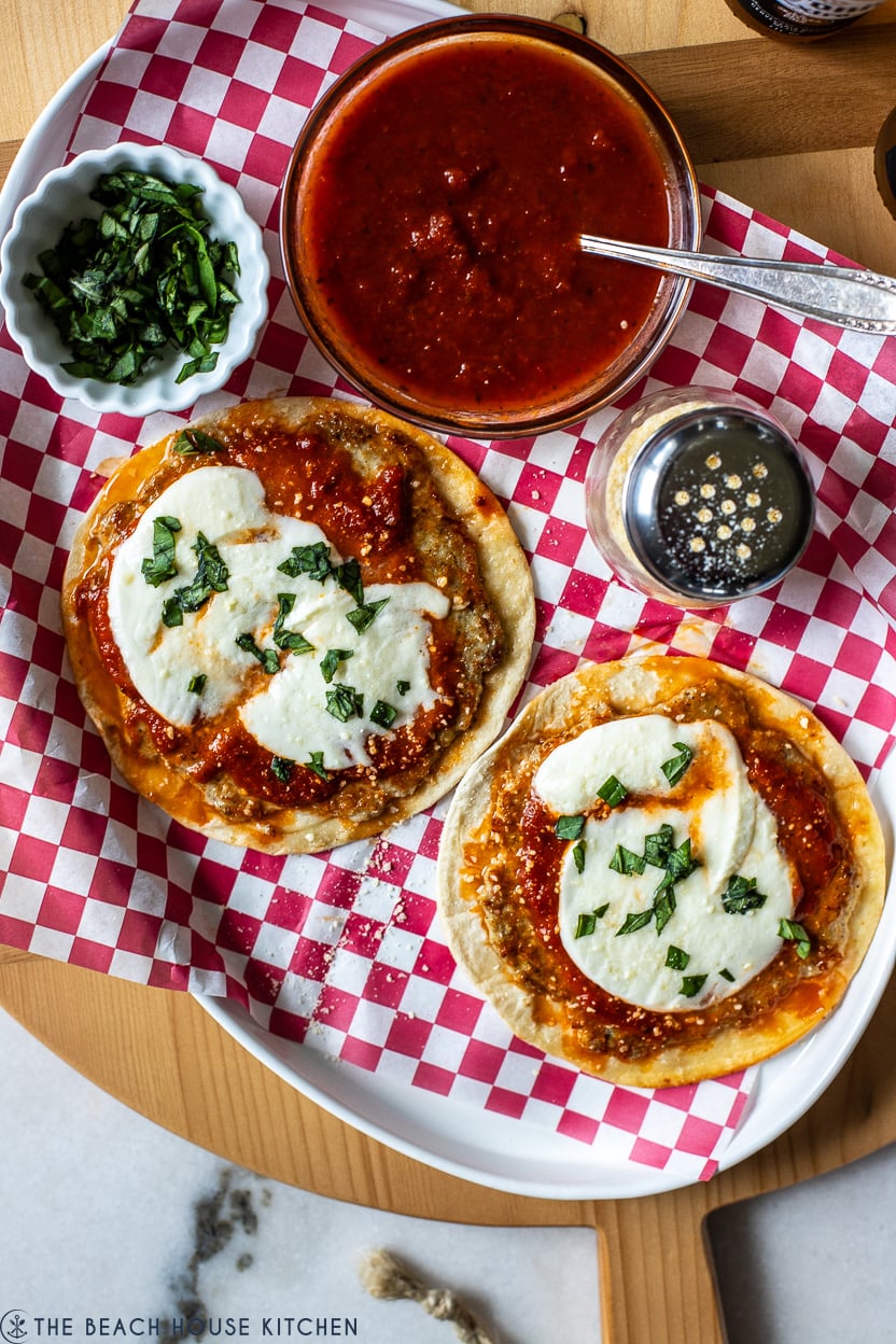 Overhead photo of two chicken parmesan smash tacos on a plate lined with red and white checked paper with a bowl of marinara and a small bowl of chopped basil.