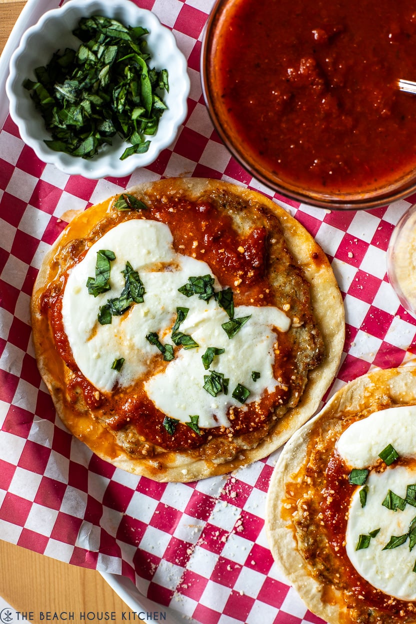 Up close overhead photo of two chicken parmesan smash tacos on a plate lined with red and white checked paper with a bowl of marinara and a small bowl of chopped basil.