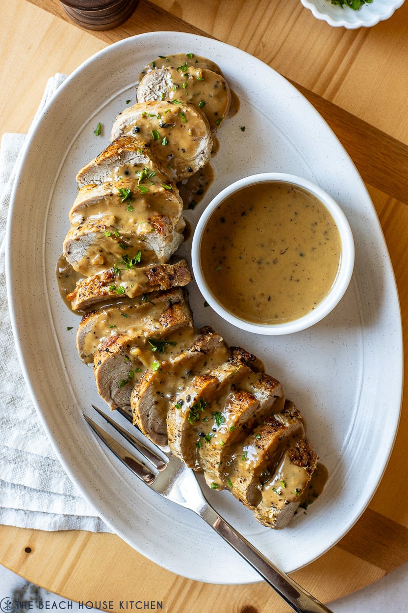 Up close overhead photo of pork tenderloin drizzled with peppercorn sauce and a small white bowl of extra sauce