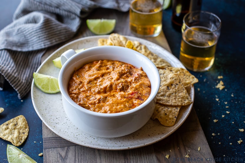Photo of a white bowl of cheesy dip on a plate with torilla chips and lime wedges