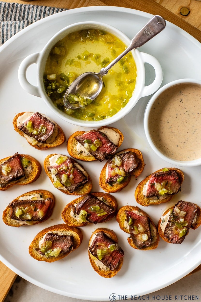 Overhead photo of a white platter of steak crostini bites and a small dish of buttery green onions