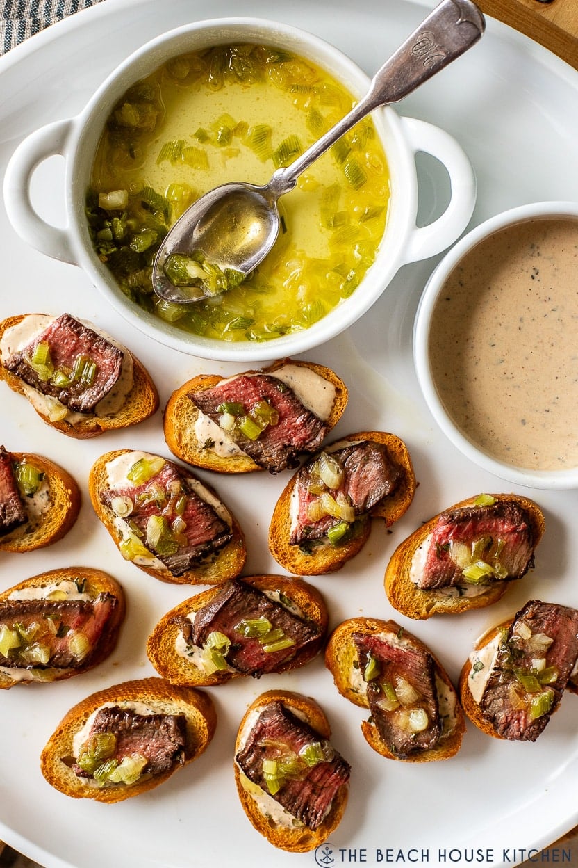 Up close overhead photo of a white platter of steak crostini bites with a small dish of buttered green onions