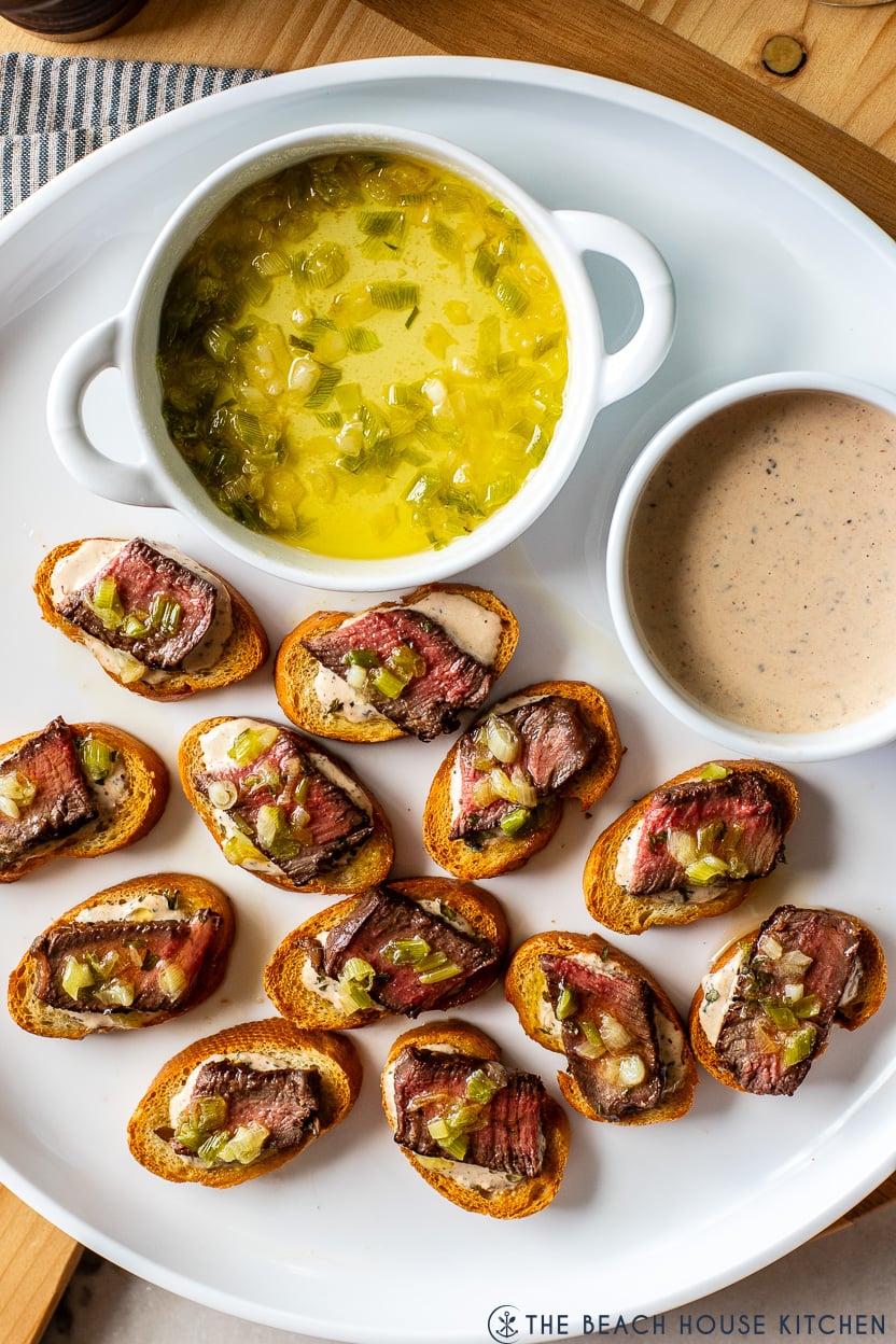 Overhead photo of a white platter of steak crostini bites with some green onions in butter