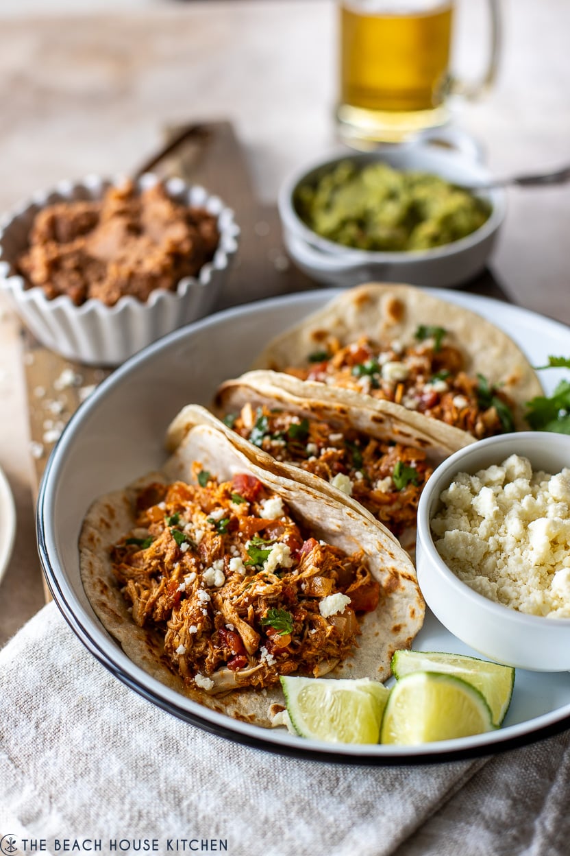 Photo of rotisserie chicken tacos in a round dish with limes wedges and and small bowl of crumbled cheese
