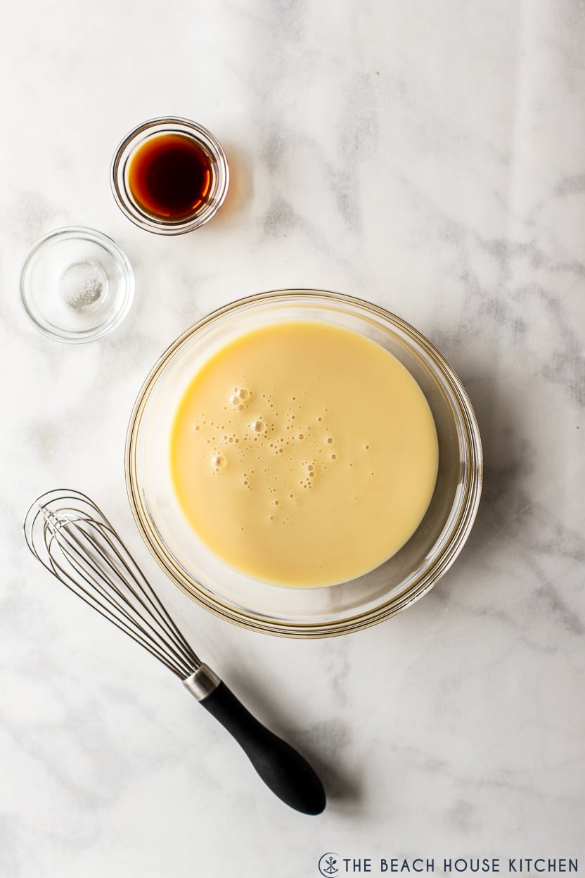 Overhead photo of a bowl of sweetened condensed milk, a small bowl of vanilla extract and salt and a whisk