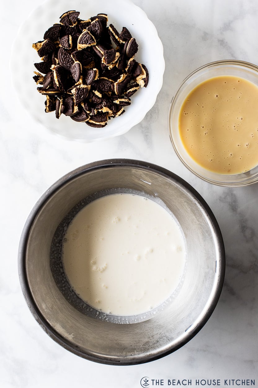 Overhead photo of a silver bowl filled with heavy cream, a bowl of chopped Oreos and a bowl of sweetened condensed milk
