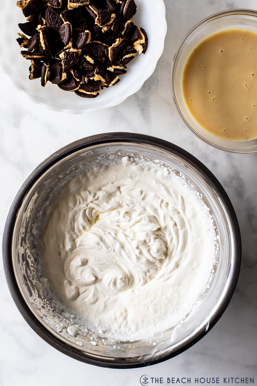 Overhead photo of a silver bowl of whipped cream, a bowl of chopped Oreos and a bowl of sweetened condensed milk