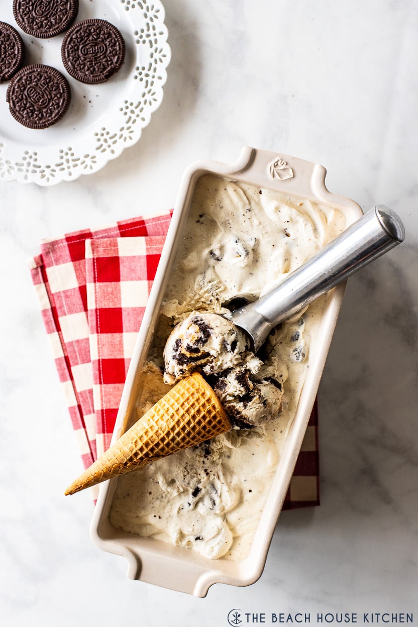 Overhead photo of a loaf dish filled with no churn oreo ice cream, a sugar cone and a silver scoop with a plate of oreos off to the side
