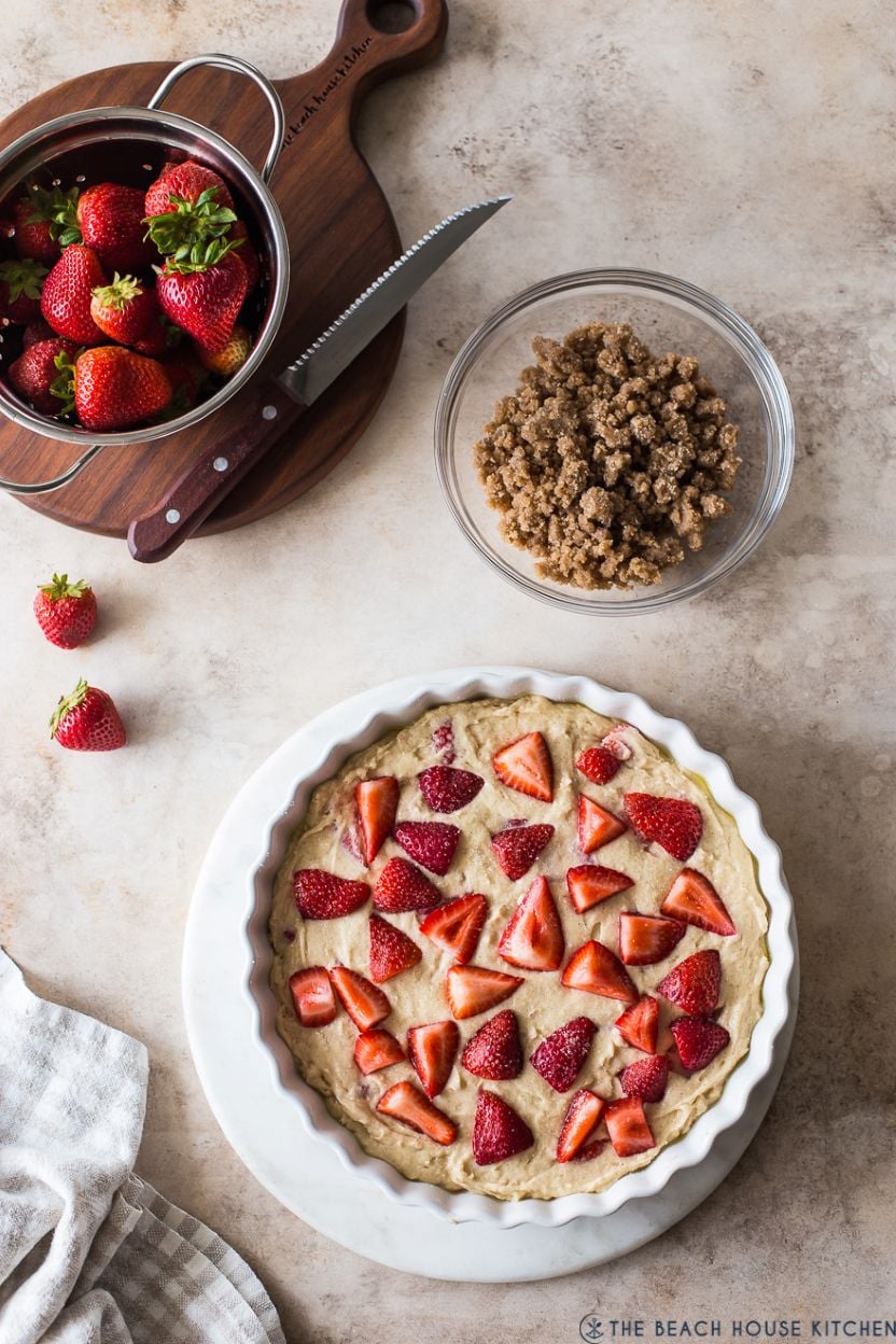 Overhead photo of a pre-baked strawberry buckle, a bowl of streusel and a bowl of strawberries