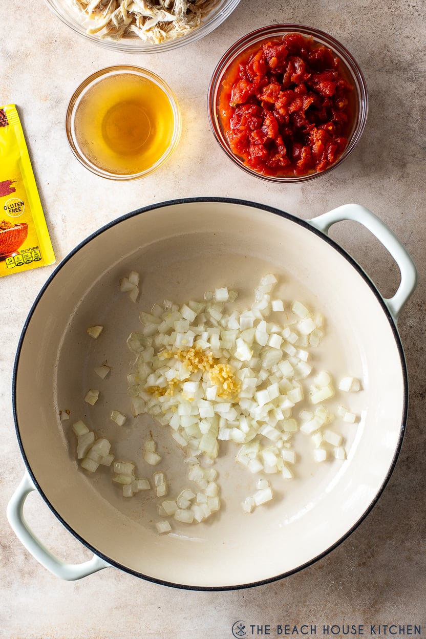Overhead photo of a skillet filled with chopped white onions and garlic with a small bowl of chopped tomatoes off to the side