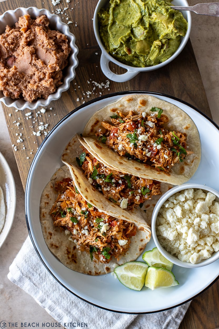 Overhead photo of a white dish filled with rotisserie chicken tacos, lime wedges and a small bowl of crumbled cheese