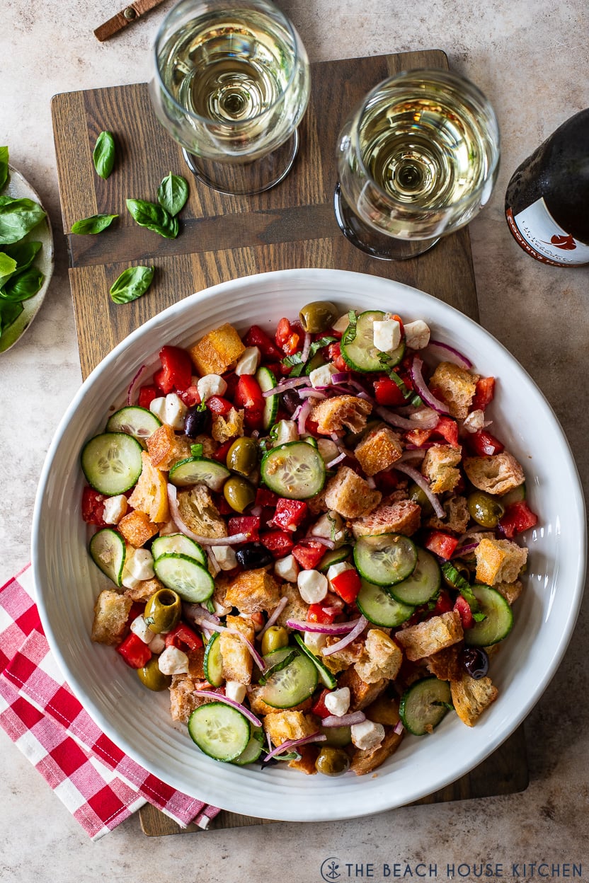 Overhead photo of panzanella salad on a wooden board with two glasses of white wine
