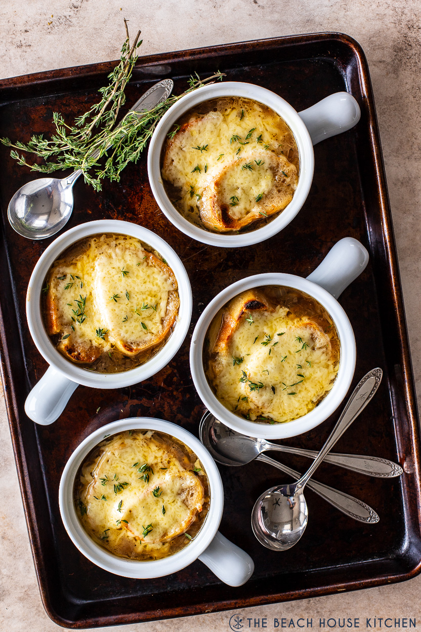 Overhead photo of four crock of French Onion soup on a baking sheet with spoons and fresh thyme sprigs