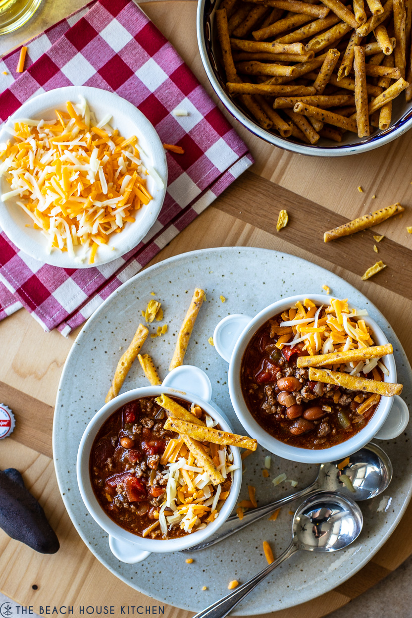 Overhead photo of two bowls of beef enchilada chili topped with shredded cheese on a plate with two spoons