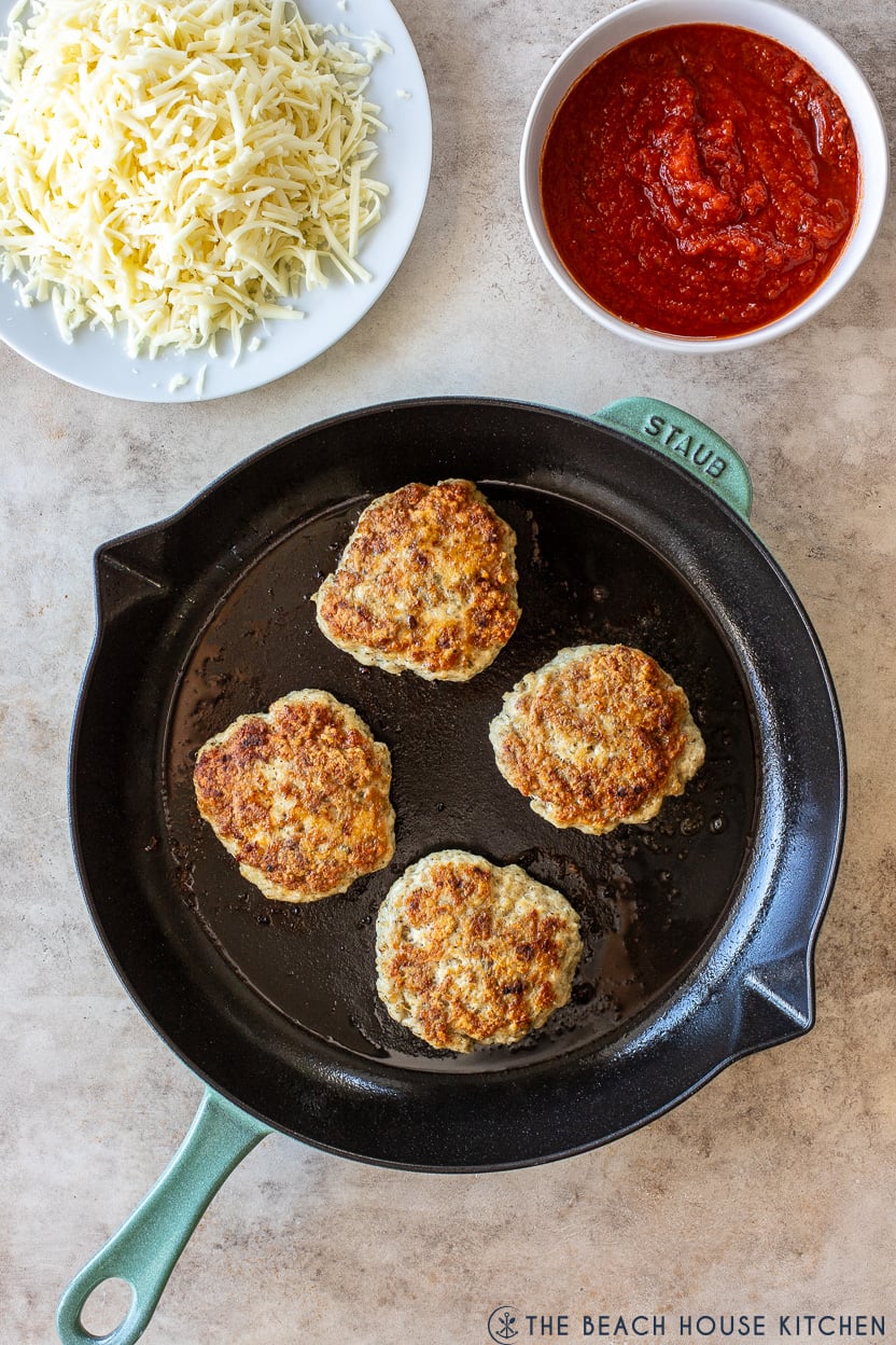Overhead photo of cooked chicken burgers in a skillet with a plate of shredded cheese and a bowl of tomato sauce off to the side