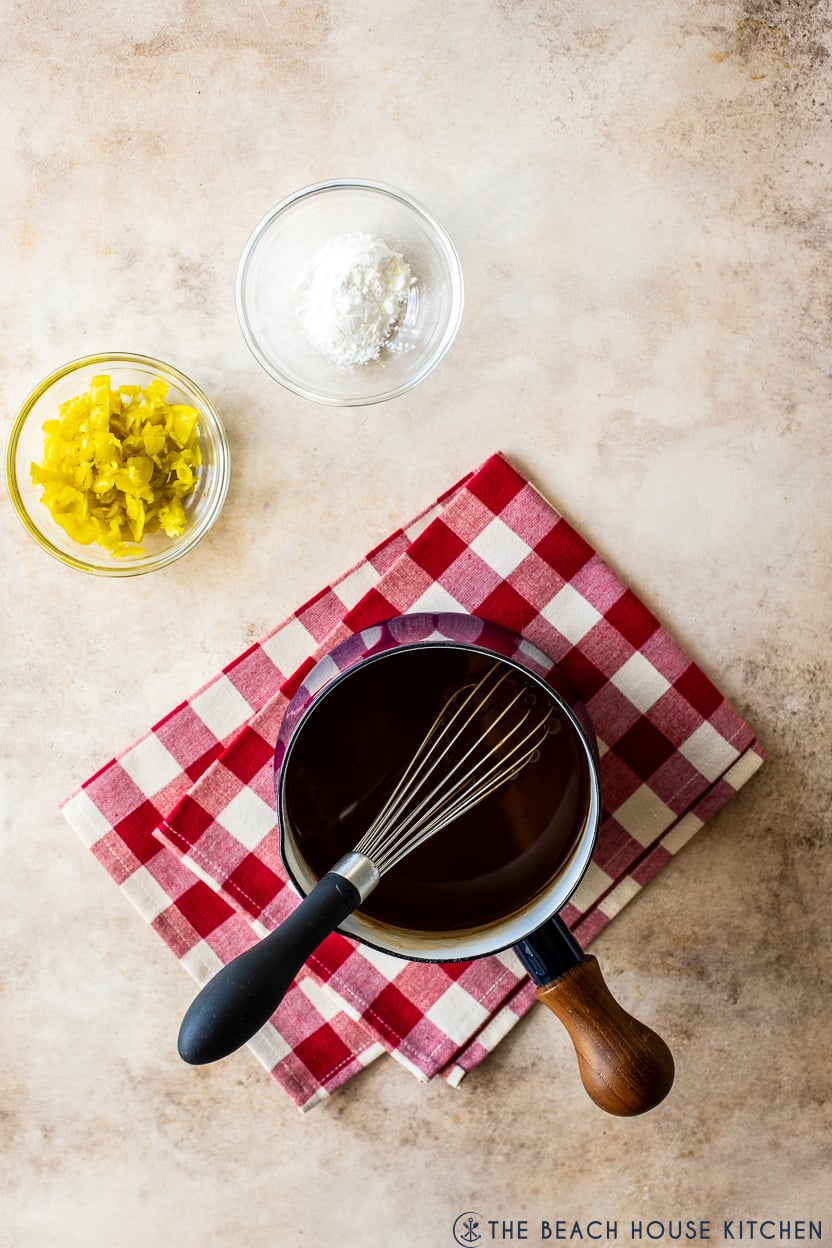 Overhead photo of a small pot of au jus gravy on a red and white checked napkin with a small bowl of peppers and a small bowl of cornstarch