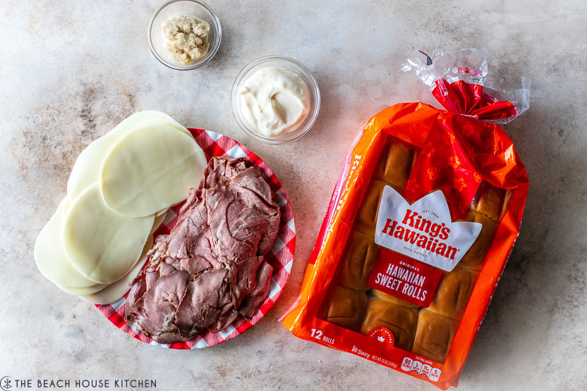Overhead photo of a plate of beef and provolone cheese slices, a bag of Hawaiian rolls and a small bowl of horseradish and mayo