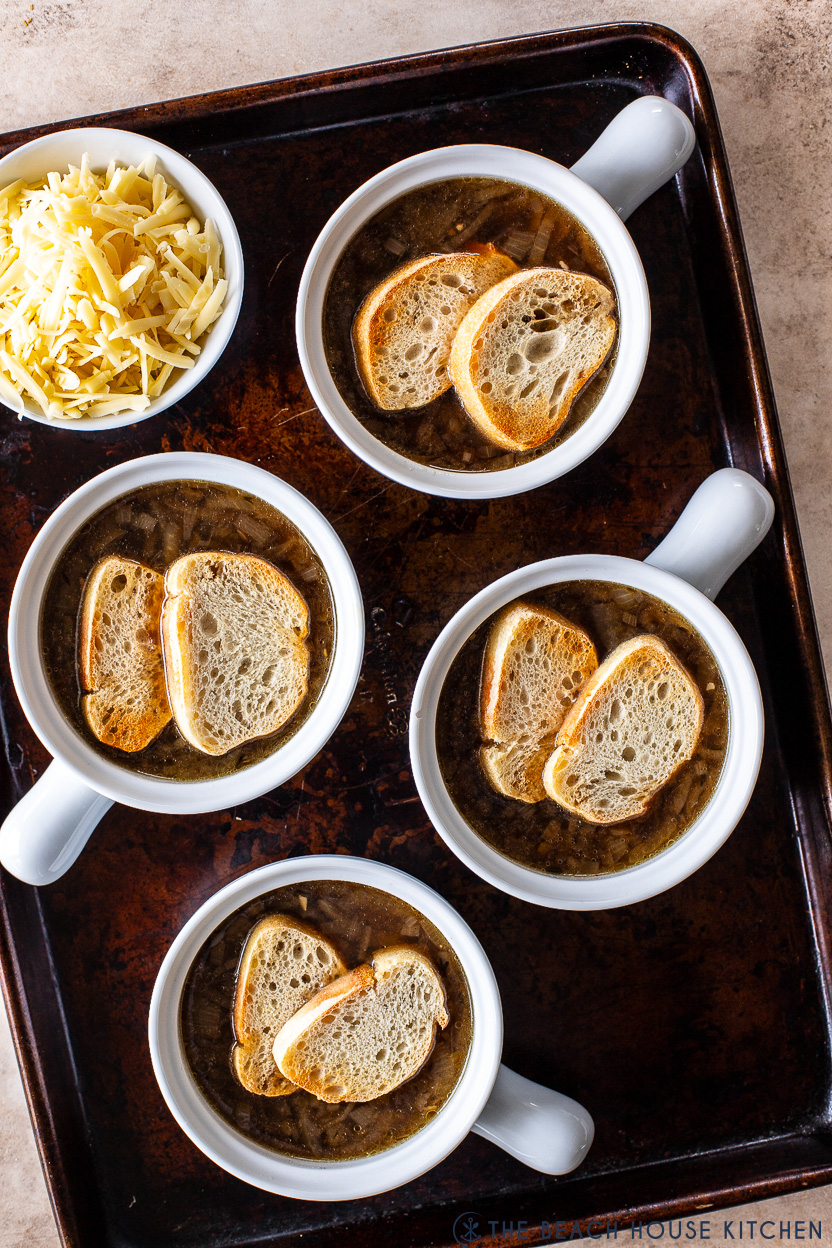 Overhead photo of four cracks of French onion soup topped with baguette slices with a bowl of shredded cheese off to the side